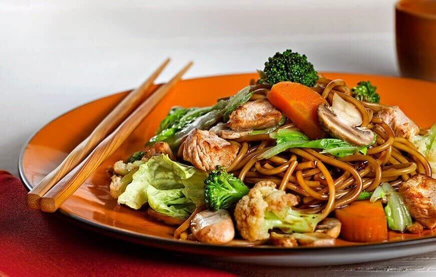 Prato de macarrão ao estilo oriental com frango e legumes, como brócolis, cenoura, cogumelos e couve-flor, servido em prato laranja com hashis sobre a mesa.