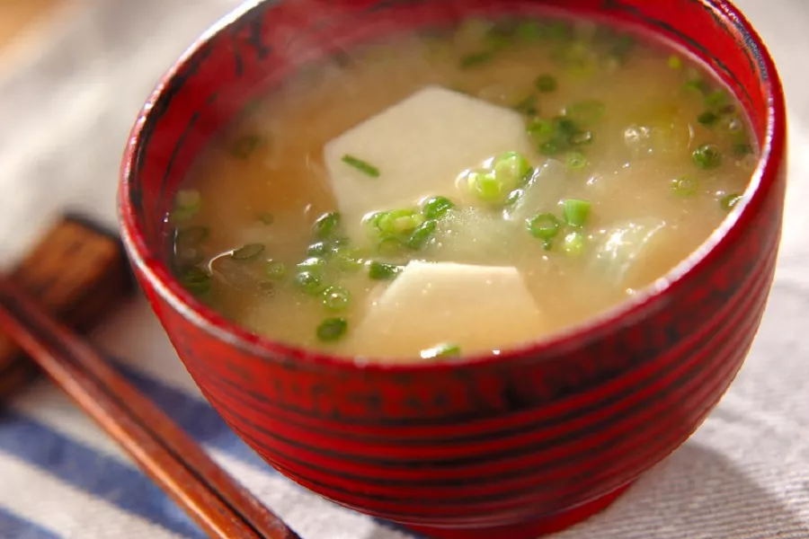 Sopa em tigela vermelha com caldo claro e pedaços de tofu, finalizada com cebolinha picada e servida com hashi sobre tecido claro. Missoshiru