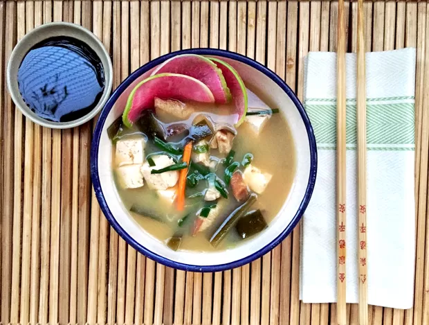Missoshiru. Tigela de caldo com legumes e tofu, decorada com fatias de cebola roxa e ervas, ao lado de guardanapos e hashis sobre esteira de bambu. Vista de cima.
