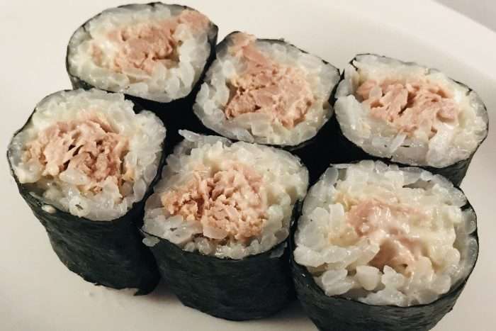 Nove pedaços de hossomaki com arroz ao centro e recheio de atum e maionese, preparados e cortados em rolinhos, em uma travessa branca.