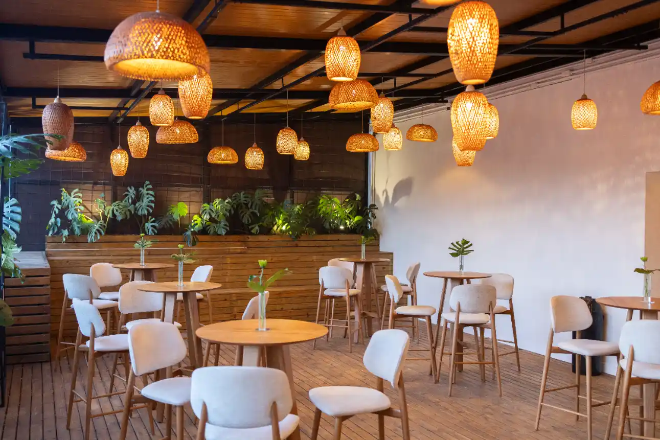 Ambiente do Espaço V com mesas e cadeiras de madeira e cadeiras estofadas brancas, decoração com plantas em floreiras e lustres pendentes de iluminação dourada