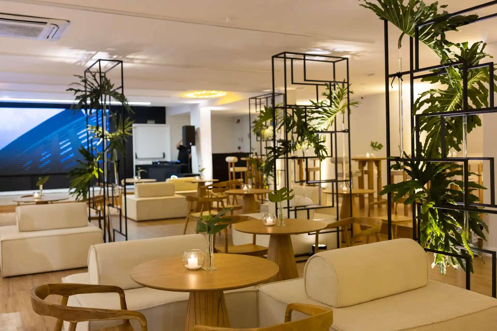 Ambiente interno de do Espaço V moderno com mobiliário claro, mesas redondas, poltronas, plantas em vasos e uma área de convivência iluminada, com painéis e estrutura metálica decorativa