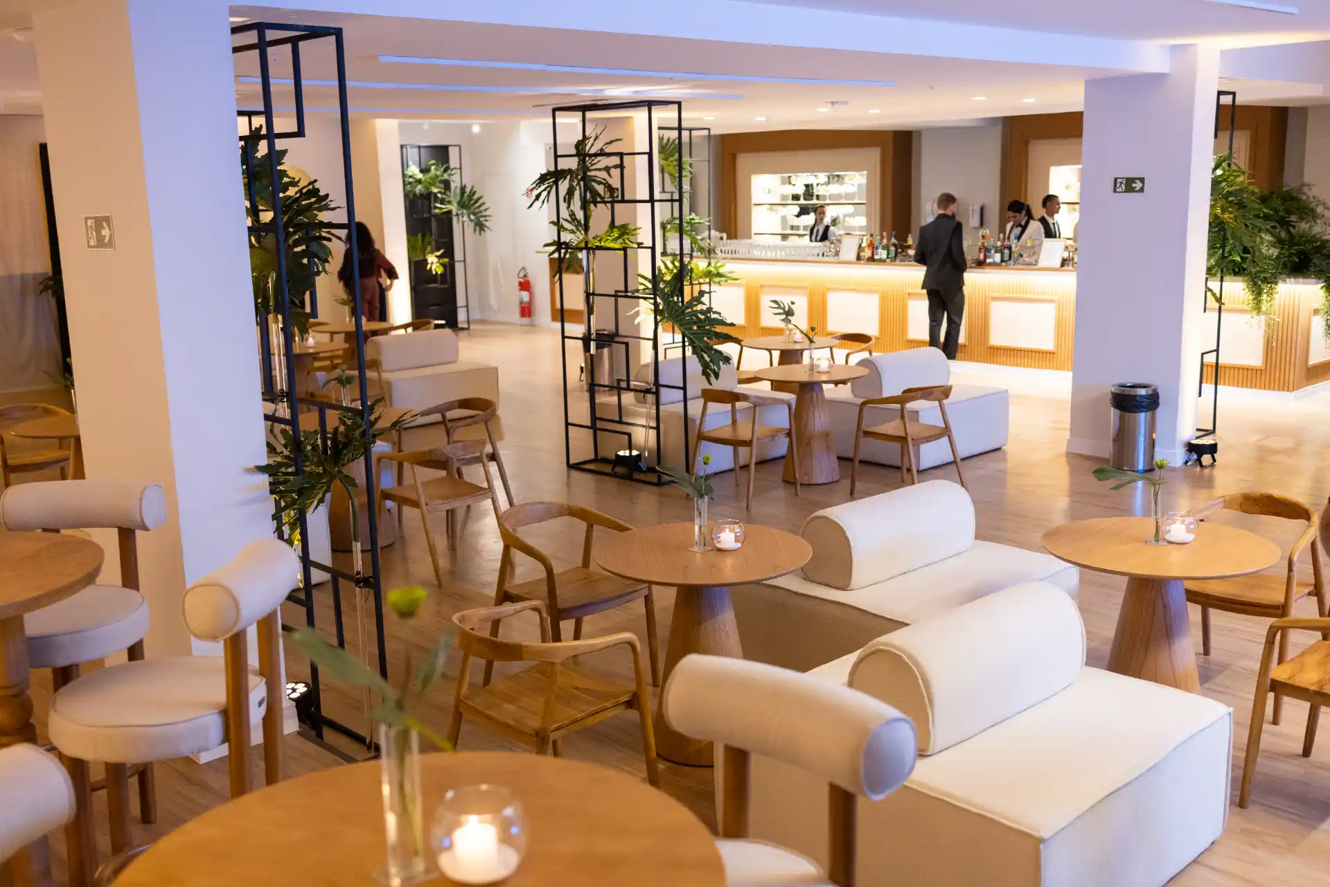 Ambiente de lounge do Espaço V com mesas redondas, assentos estofados claros, decoração com plantas e iluminação indireta, vista de uma área de recepção e bar ao fundo.