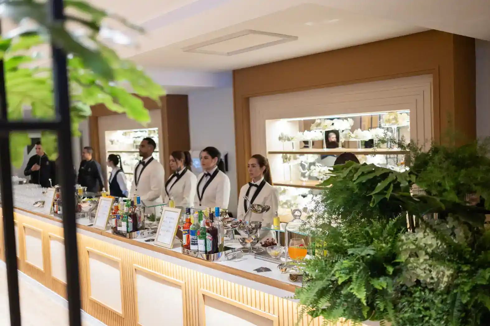 Equipe uniformizada serve bebidas em um evento em salão com mesas de coquetel, decoração floral e copos sobre o balcão