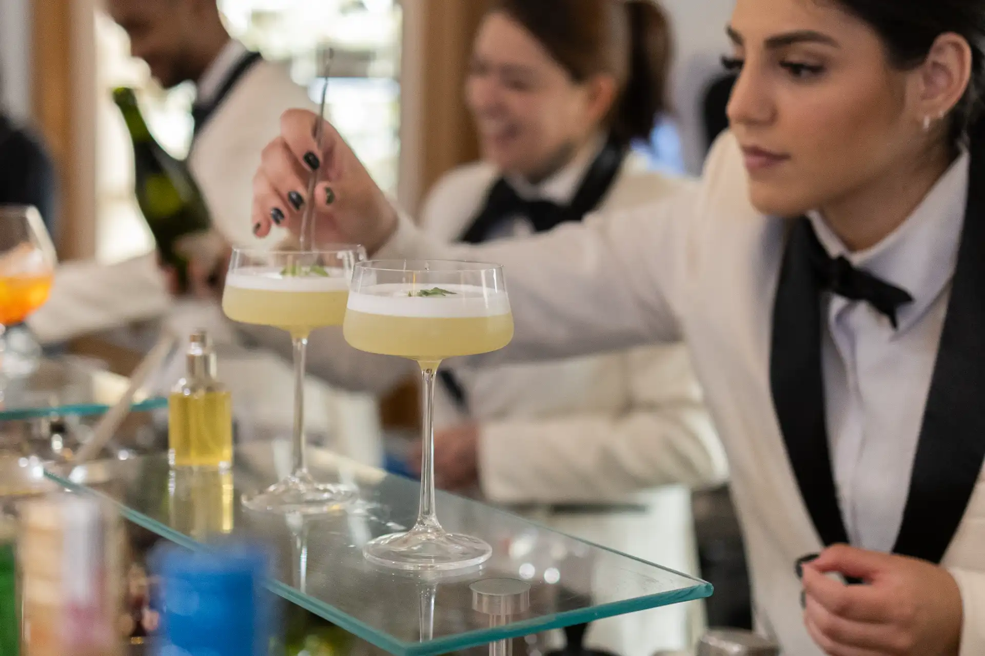 Garçons em traje social servindo drinques em taças com diferentes cores, em um evento requintado, com foco no atendimento à mesa