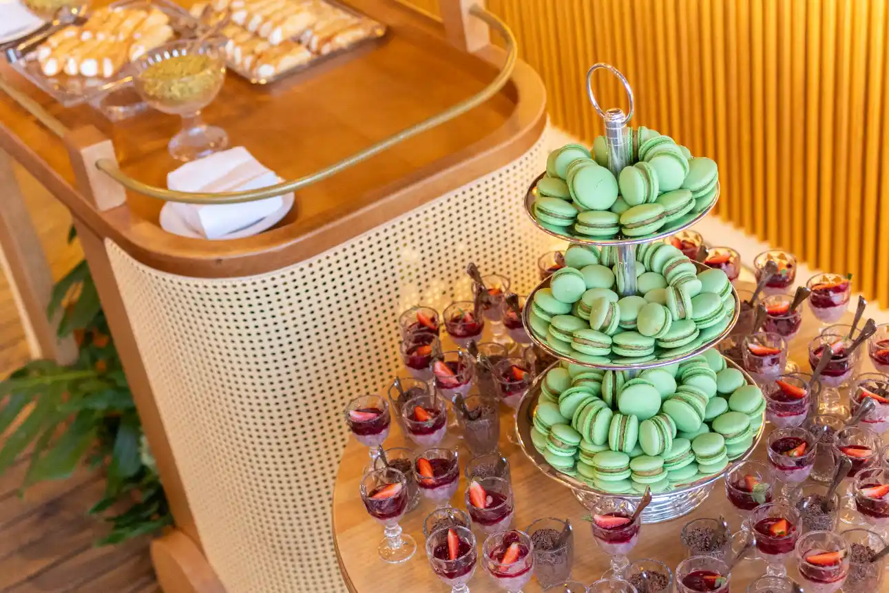 Mesa de festa com torre de macarons verdes em camadas e mini taças decorativas com bebida, em ambiente elegante e iluminado, ideal para evento ou coquetel.