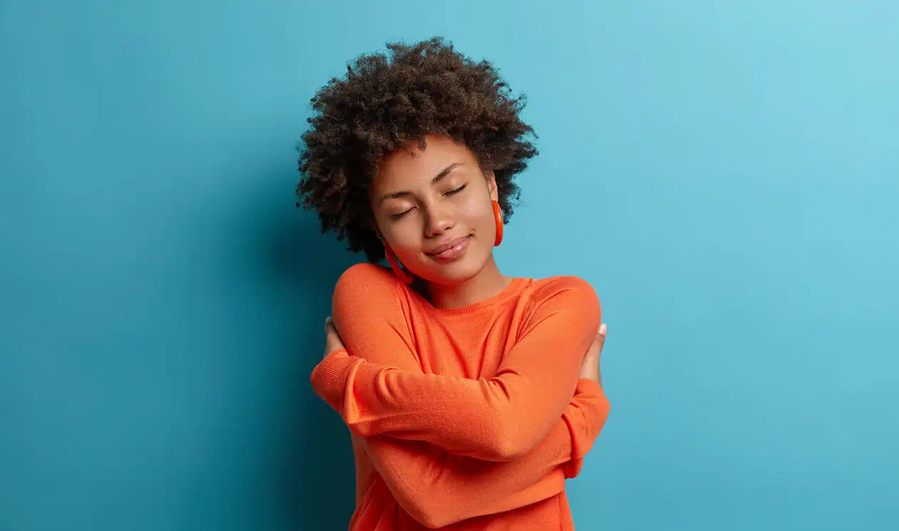 Mulher jovem com cabelo crespo usando fones de ouvido coloridos, vestindo suéter laranja e braços cruzados, com expressão calma, em fundo azul claro. autocuidado