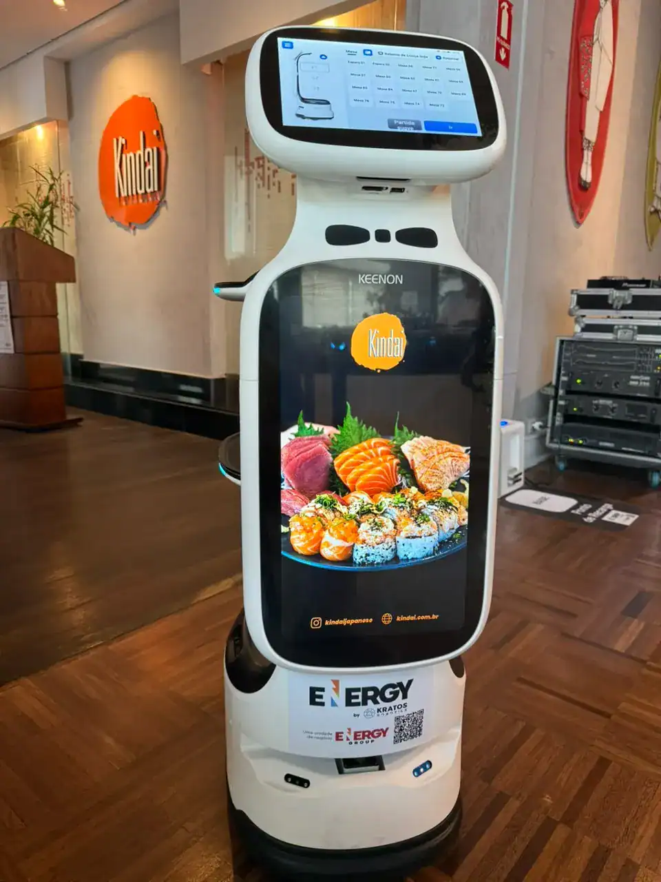 Robô de atendimento em loja, com tela de interface exibindo opções e ilustração de sushi ou peixe no visor, ao lado de equipamentos de som e decoração industrial. Robô garçom - restaurantes Kindai, Bellini e Vick