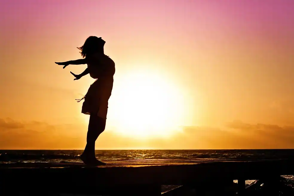 Silhueta de uma mulher correndo na praia ao pôr do sol, com o horizonte e o céu em tons quentes de rosa e laranja, criando clima de movimento e liberdade.