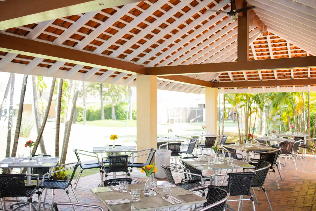 Área externa para eventos com mesa posta, cadeiras de metal e decoração com flores, sob pergolado de madeira e cobertura, em ambiente amplo com jardim ao fundo.