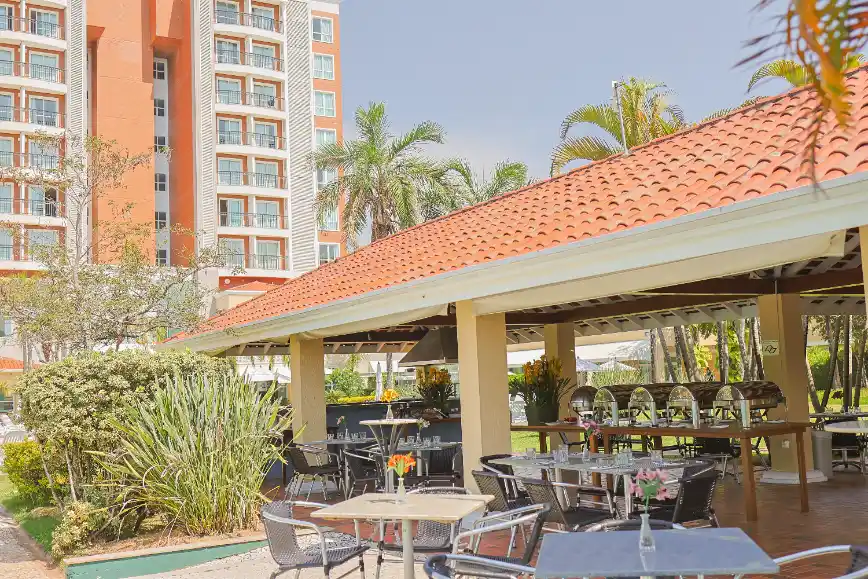 Área externa de hotel com mesas e cadeiras sob cobertura, churrasqueira, jardim com plantas e ao fundo prédio de apartamentos com varandas, em ambiente ensolarado.