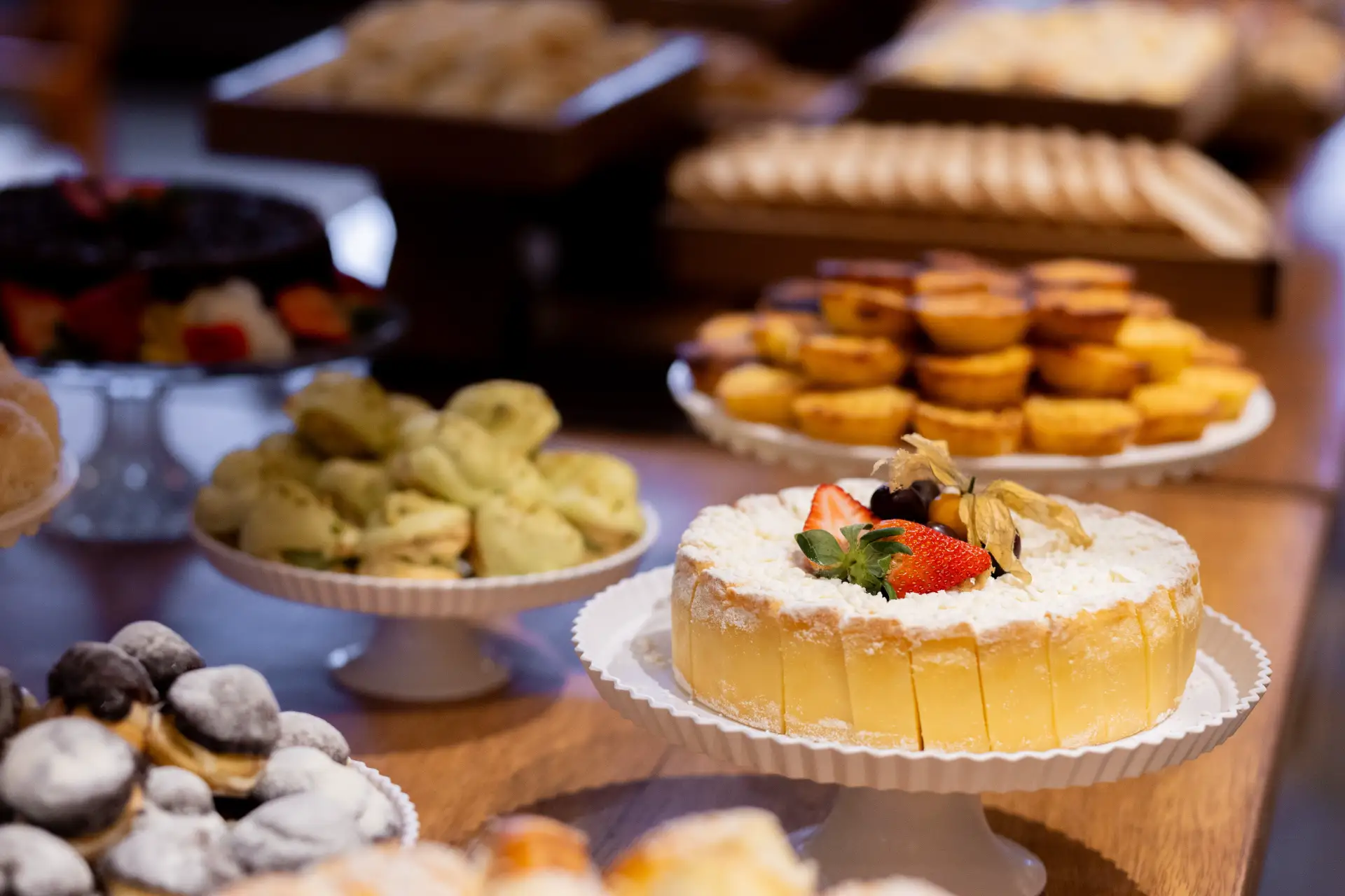 Bolo individual estilo cheesecake em uma bandeja de confeitaria, com morango e pedaços de frutas sobre a cobertura, em meio a outras sobremesas e doces exibidos no balcão.