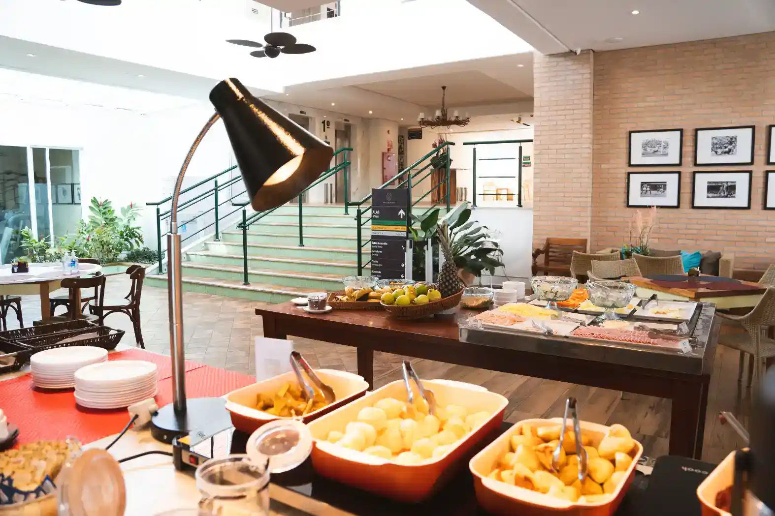 Buffet de café da manhã em ambiente interno moderno, com mesa posta e travessas com frutas, pães e alimentos, iluminação natural e decoração com plantas.