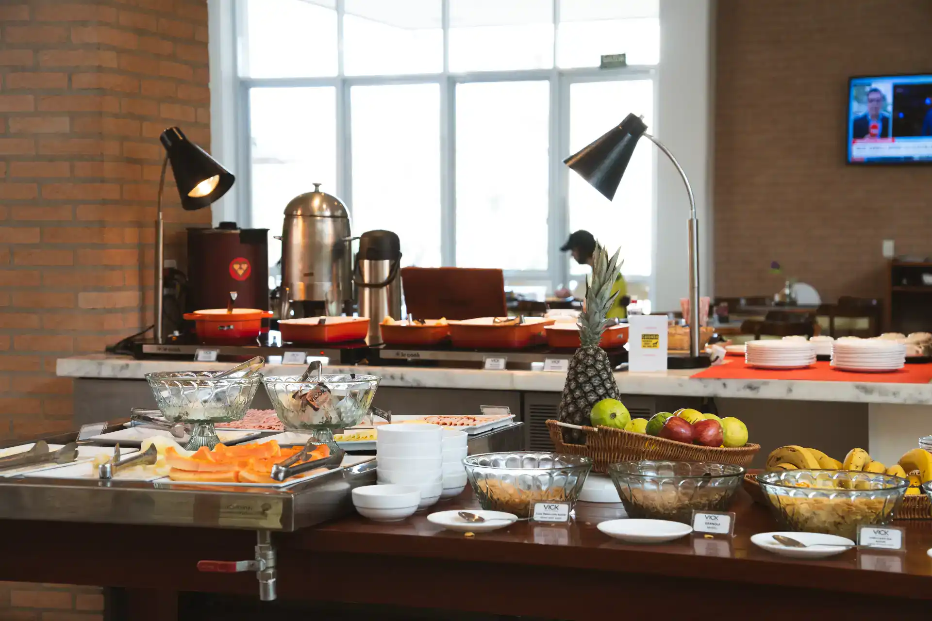 Buffet de café da manhã em hotel com mesas expostas, utensílios, frutas e sucos, copos de vidro e decoração ao fundo na sala com janelas grandes.