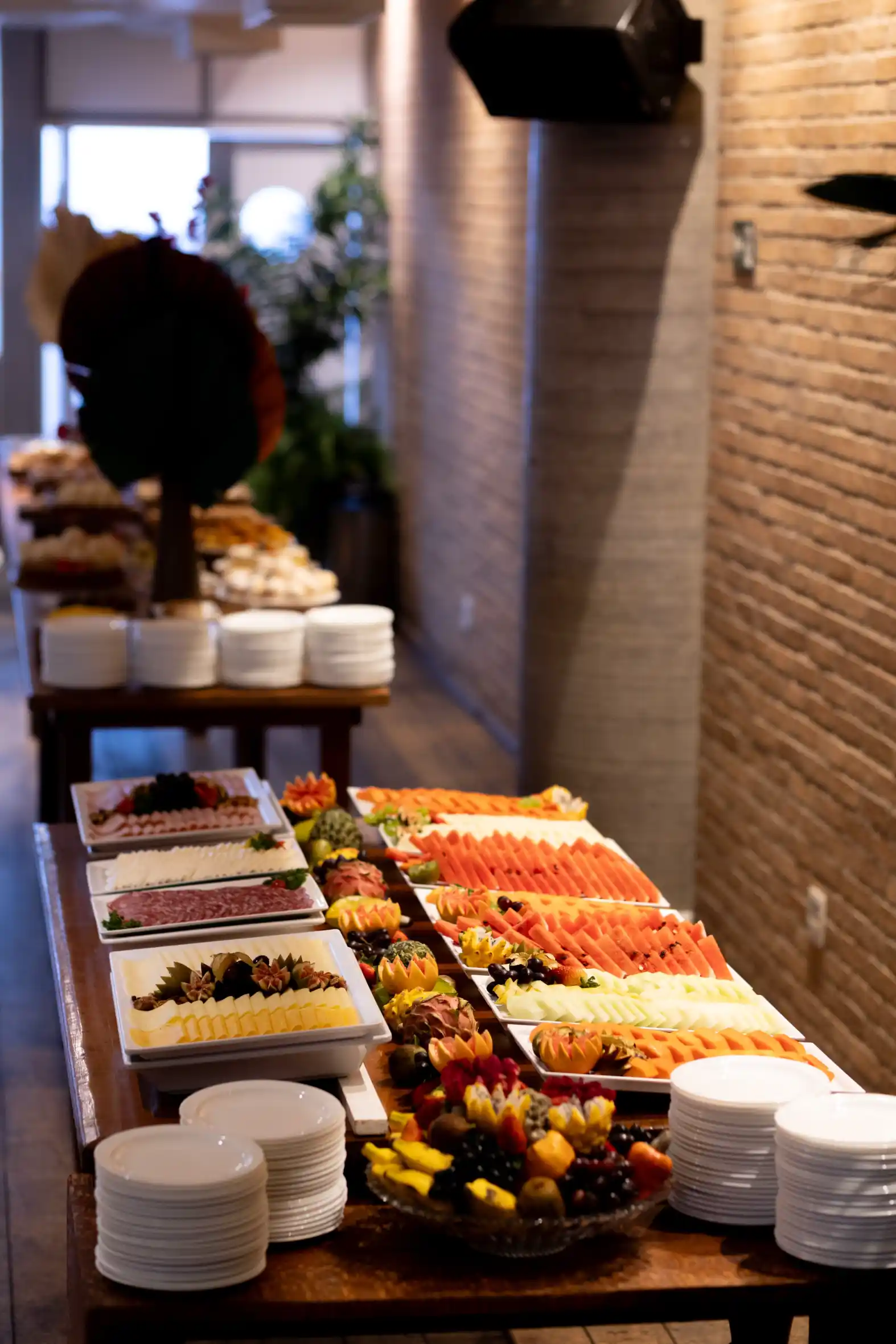 Buffet de sobremesas com frutas fatiadas e opções de chocolate em bandejas brancas, decoradas sobre balcão de madeira em ambiente com iluminação quente e parede de tijolos à vista.