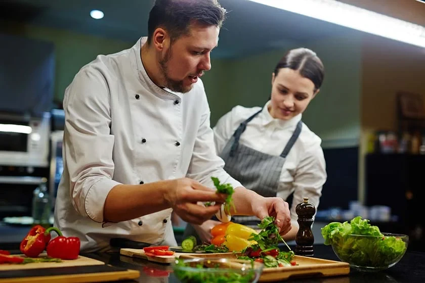 Chef preparando salada com legumes frescos em bancada de cozinha profissional, enquanto a equipe observa ao fundo. A imagem mostra verduras, pimentões e temperos sendo montados.