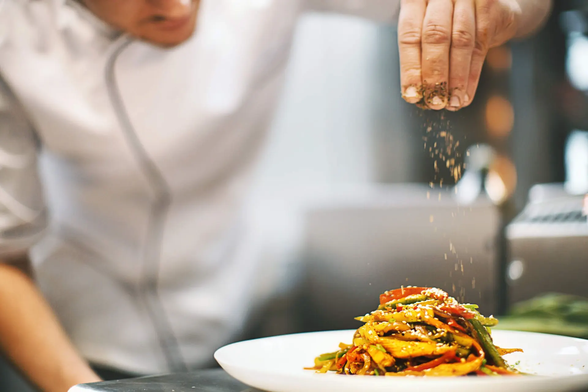 Chefe em cozinha prepara um prato com legumes e temperos enquanto ingredientes são jogados sobre a comida em uma travessa branca, com fundo desfocado.