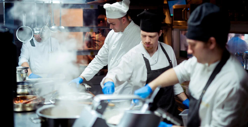 Chefs de cozinha em restaurante preparando pratos, com luvas e aventais brancos, panelas e utensílios ao fundo, em ambiente profissional de culinária