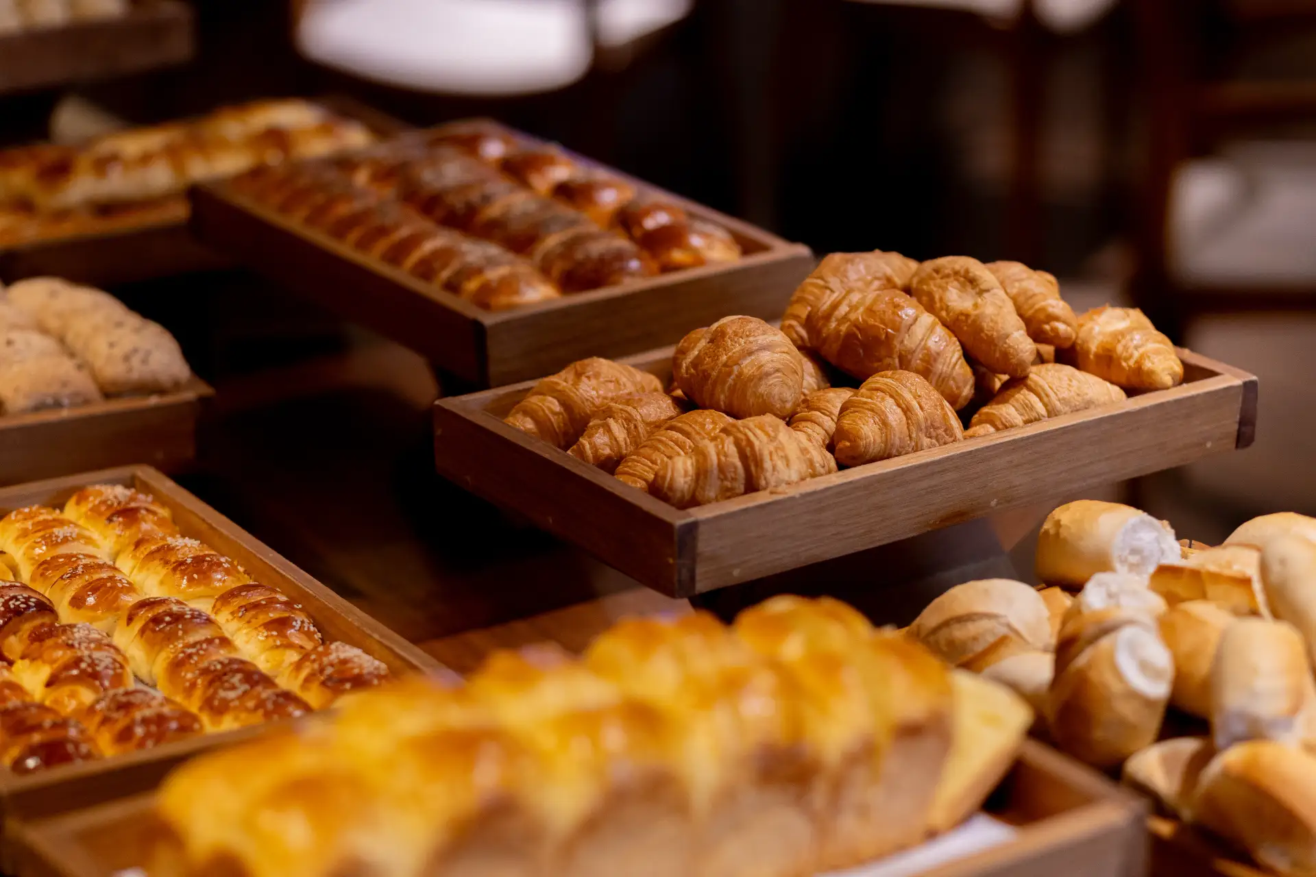 Diversas bandejas com croissants e pães doces assados em uma mesa de confeitaria, com foco em croissants dourados e folhados expostos para venda.