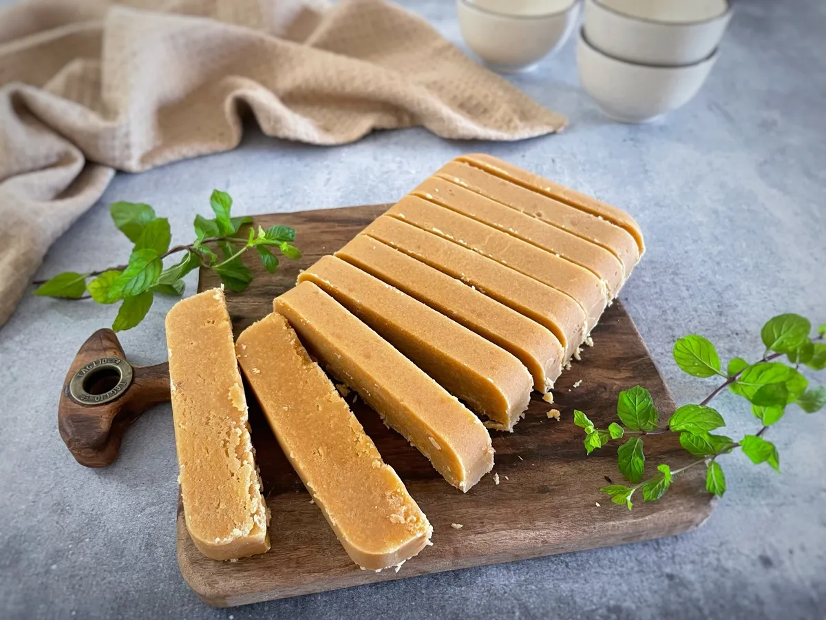 Barra de doce de leite fatiada em pedaços sobre uma tábua de madeira, com folhas verdes ao redor e pano ao fundo sobre superfície cinza, pronto para servir.