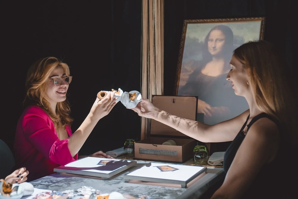 Duas mulheres em uma mesa jantando com experiências de arte, com caixas e livros abertos ao fundo; ao fundo há uma pintura clássica e clima descontraído.