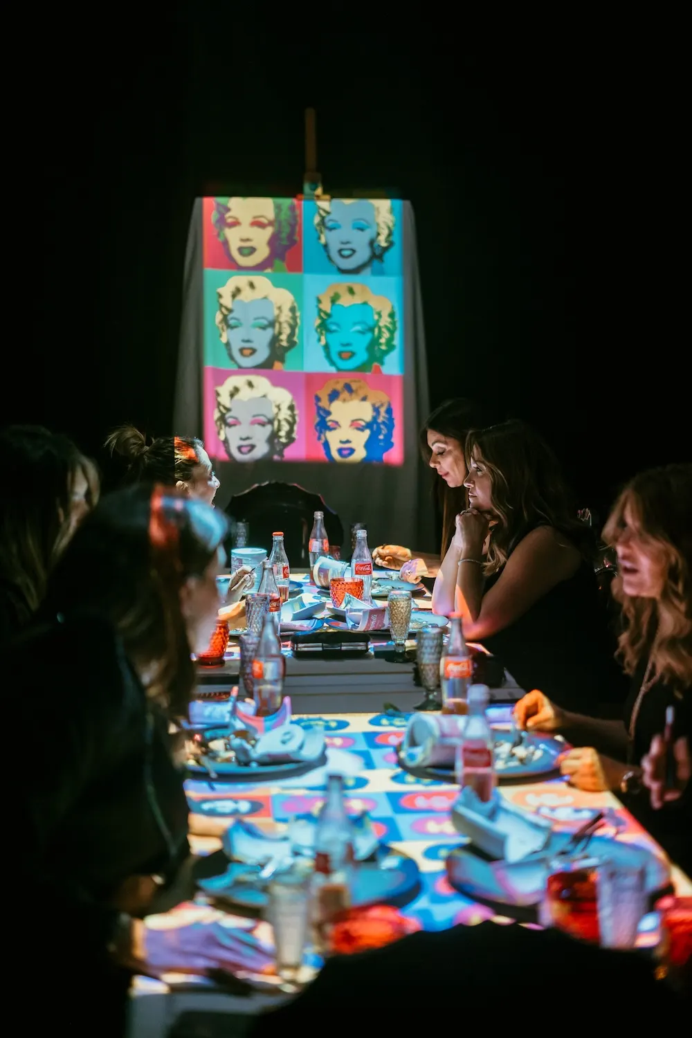 Mesa de jantar com pessoas sentadas em um evento descontraído, bebidas e pratos sobre a toalha colorida, com telão iluminado ao fundo exibindo imagens em estilo pop art