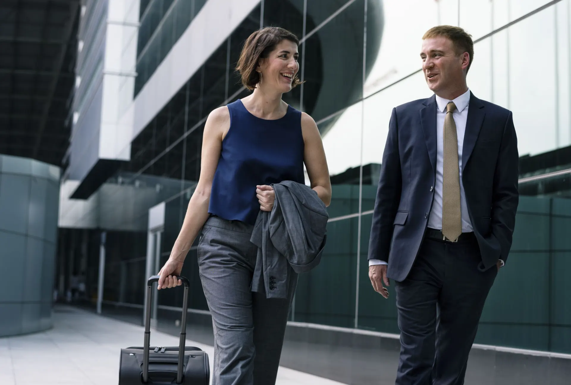 Executivos caminhando ao ar livre em um ambiente corporativo moderno, uma mulher com mala de rodinhas e casaco nas mãos, enquanto um homem de terno conversa e sorri ao lado dela.