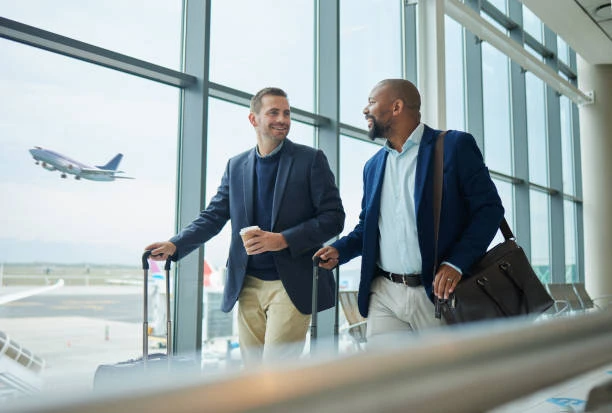 Dois homens executivos conversam e fazem check-in em um aeroporto, com malas e carrinho de bagagem, enquanto um avião decolagem aparece ao fundo através de grandes janelas.