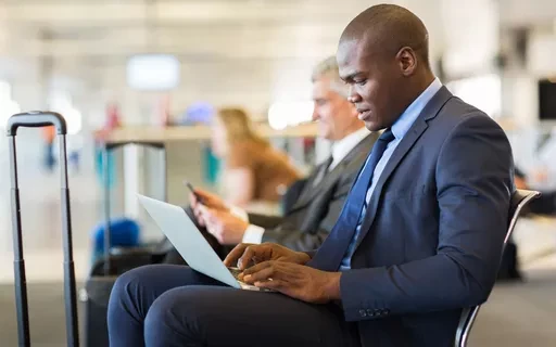 Homem de terno trabalhando em notebook em ambiente corporativo, digitando e lendo documentos enquanto aguarda ou realiza atividades no escritório.