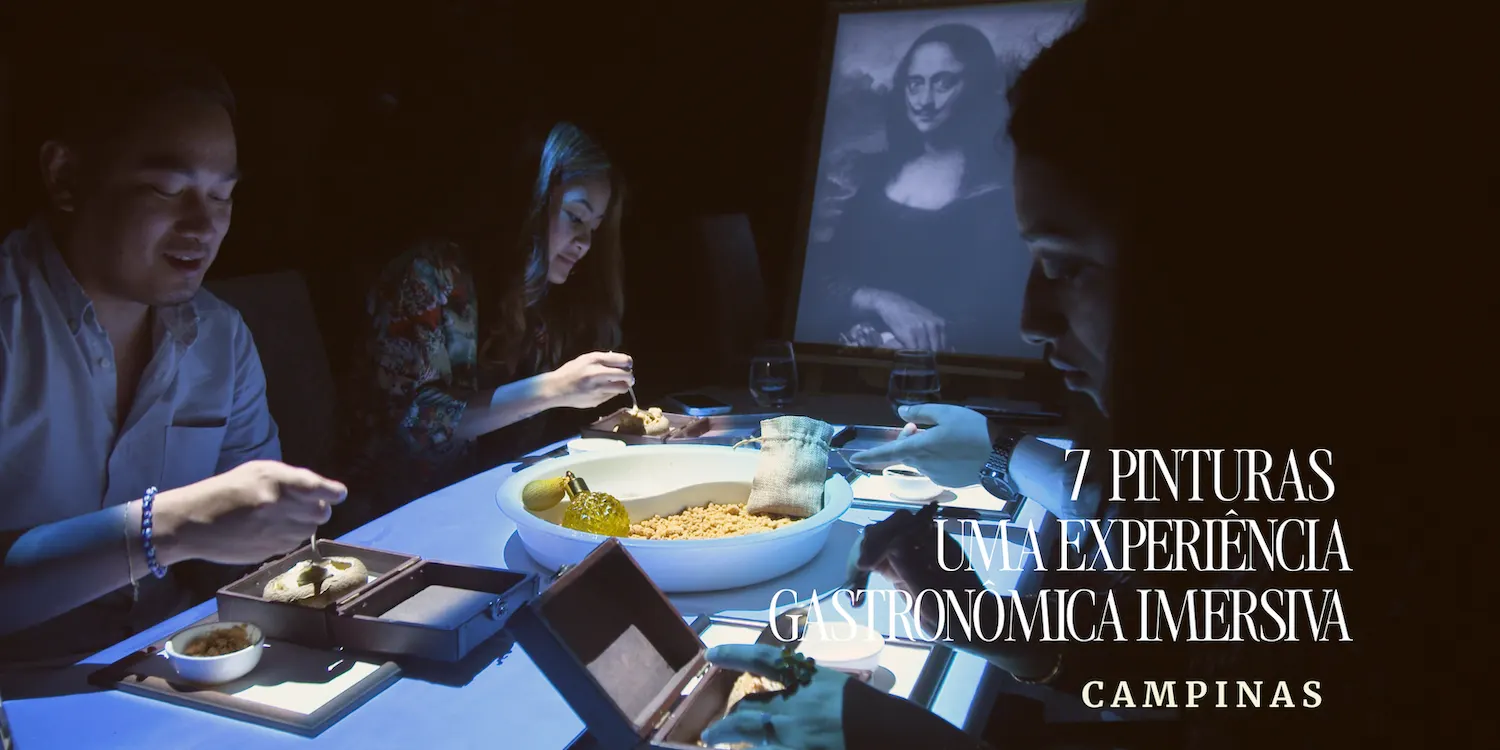 Jantar em experiência gastronômica imersiva em Campinas, com quatro pessoas ao redor de uma mesa iluminada por prato e travessa, enquanto ao fundo aparece uma imagem em tela e texto promocional “7 PINTURAS UMA EXPERIÊNCIA”.