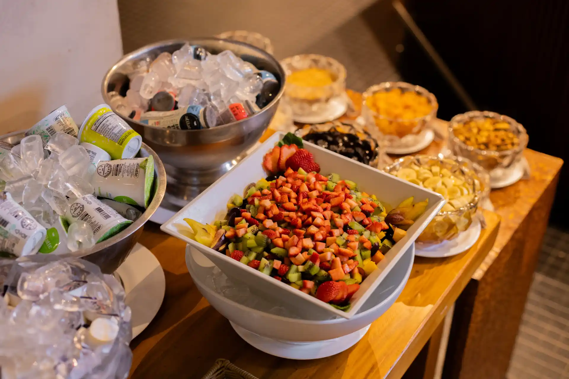 Mesa de bufê com tigela grande de frutas picadas, como morango, melão e cubos coloridos, além de recipientes com gelo, sobremesas e frutas servidas para os convidados.