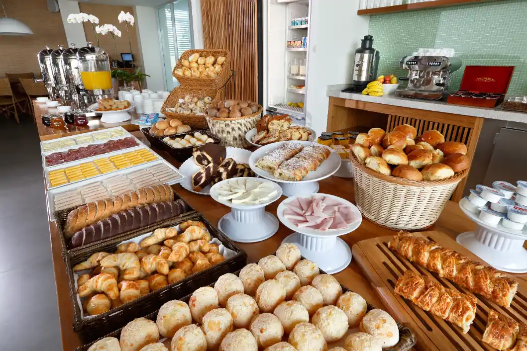 Mesa de café da manhã com diversas opções de pães, bolos e croissants em cestas e bandejas, incluindo itens doces e salgados, em um buffet.