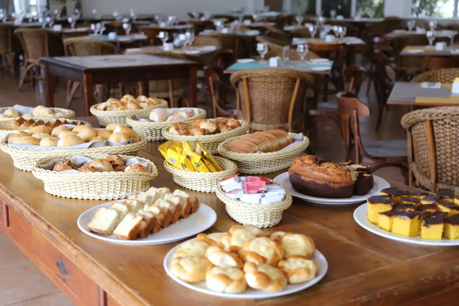 Mesa de café da manhã com pães, bolos e salgados em cestas e travessas, ao ar livre, com cadeiras ao fundo em um ambiente de restaurante ou evento.