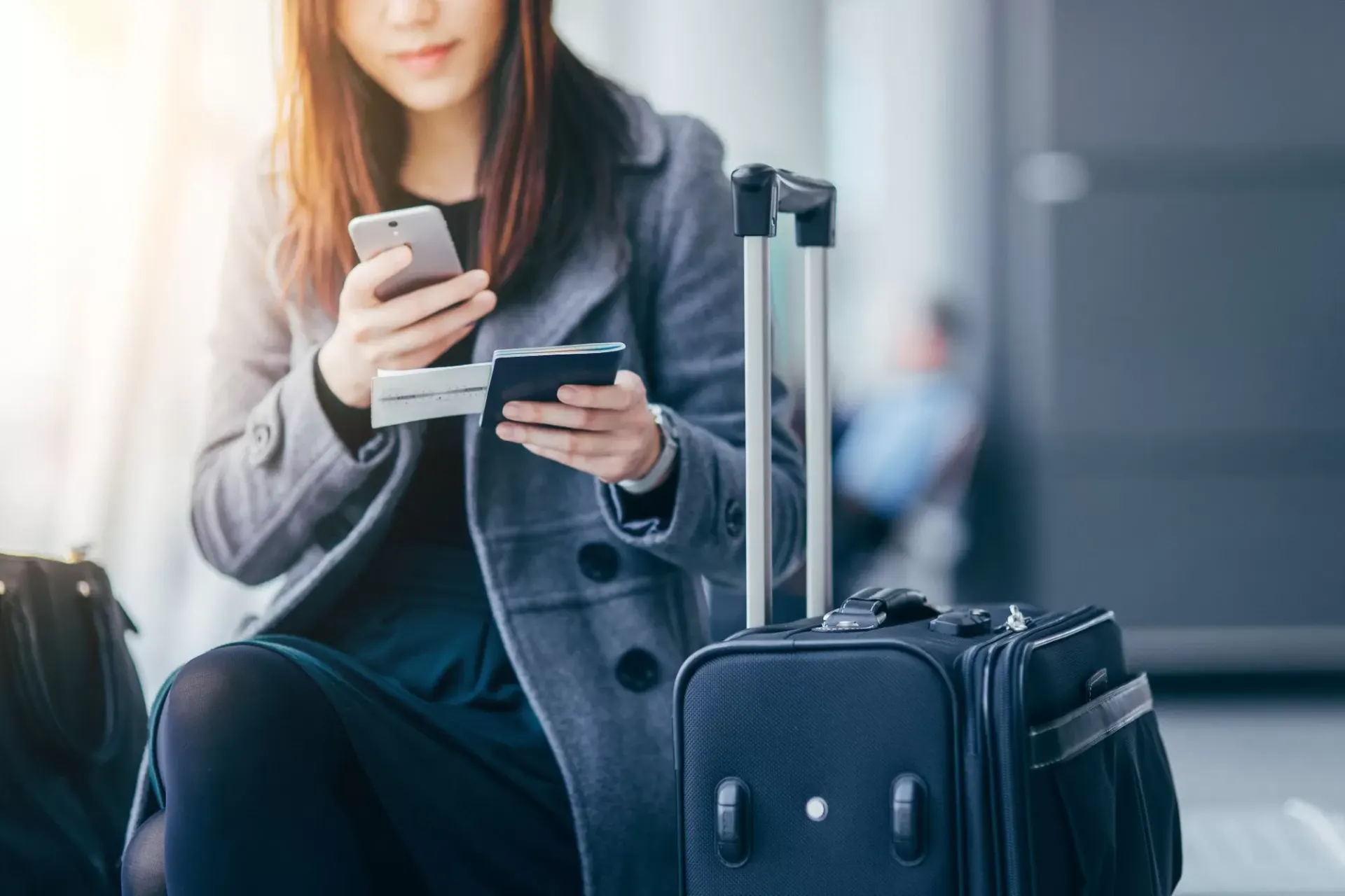 Mulher sentada no aeroporto segura celular e cartões de crédito, enquanto uma mala de viagem rígida azul está ao lado. Foto com foco em planejamento de compras e itinerário.