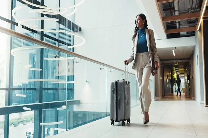 Mulher viajante com casaco claro e blusa azul, caminhando em um corredor moderno de aeroporto com mala de rodinhas, iluminação natural e arquitetura contemporânea ao fundo.