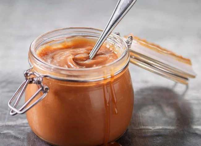 Pote de vidro com doce de leite cremoso e colher de metal servindo o creme, com fundo neutro desfocado e destaque para a textura caramelizada.