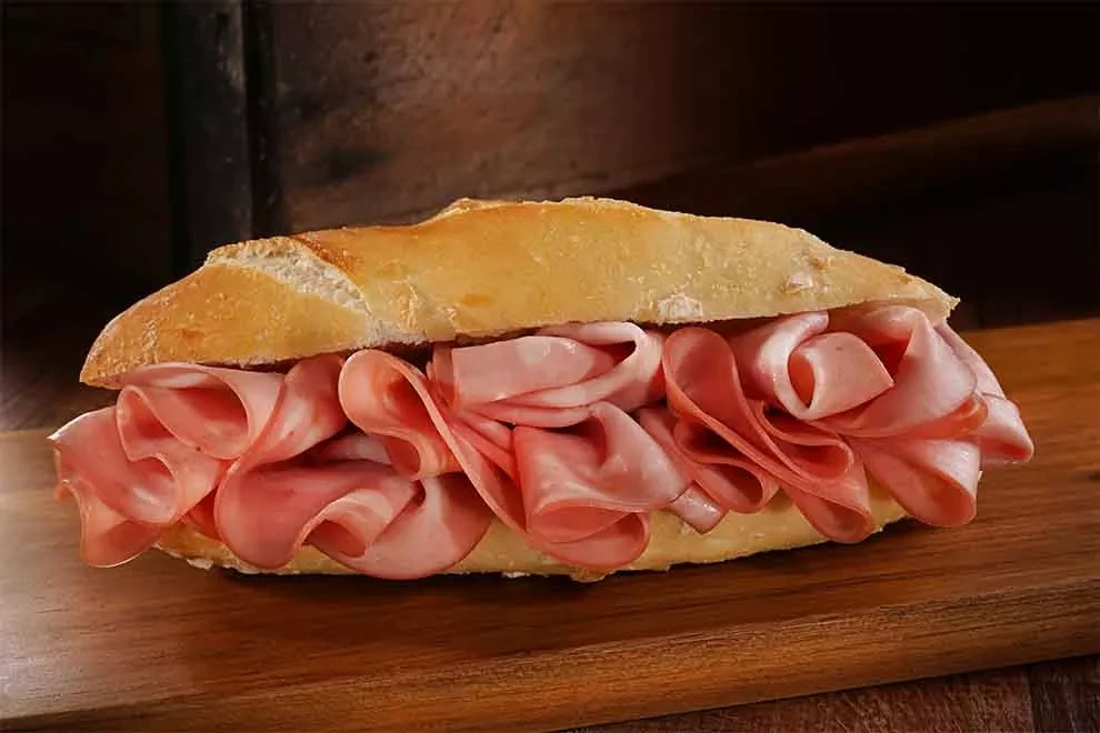 Sanduíche de pão com fatias de mortadela, com camadas de carne fatiada visíveis e suculentas sobre uma tábua de madeira.