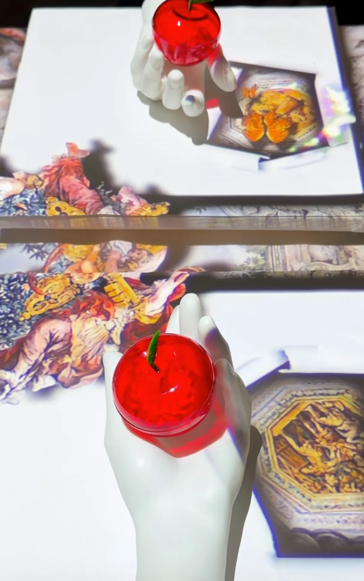 Mãos brancas com uma vela em forma de gota vermelha no centro, com sombras e iluminação forte, sobrepostas a cartões decorativos com ilustrações coloridas. Jantar que une gastronomia e arte
